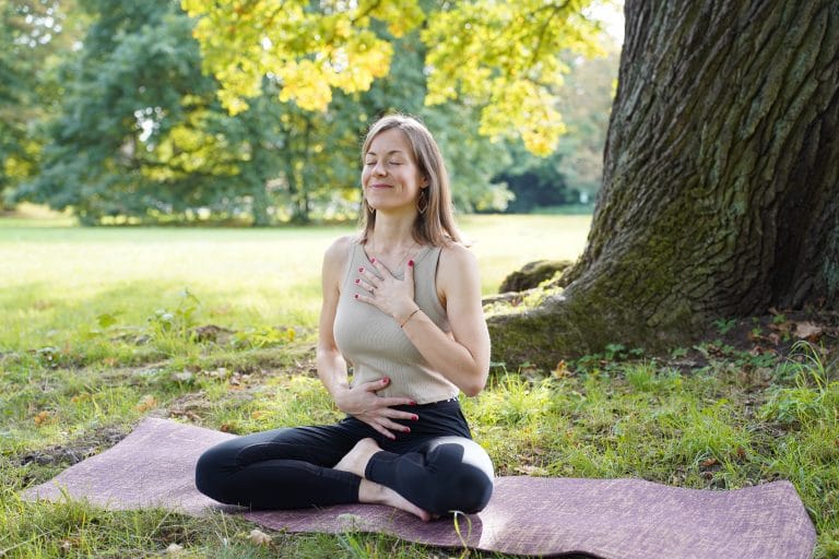 Junge Frau meditiert in der Natur. Rechts von ihr ist ein großer Baum. Die rechte Hand spürt die Atmung auf dem Bauch, die linke auf dem Brustkorb. Sonnenstrahlen kommen von hinten