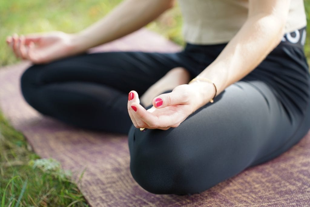 Eine Detailaufnahme von einer jungen Frau beim meditieren. Man sieht nur die geöffneten Hände auf dem Schneidersitz. Sie sitzt auf einer Yogamatte auf einer Wiese.