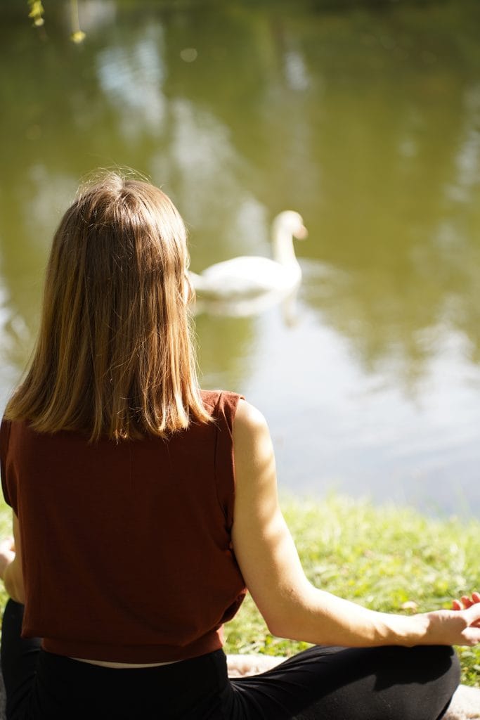 Eine junge Frau ist von hinten zu sehen, wie sie in der Natur meditiert. Vor ihr ist ein Schwan auf einem See. Die Sonne strahlt auf den See und die Frau.