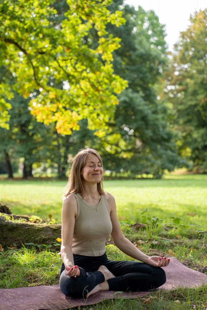 Junge Frau meditiert in der Natur. Rechts von ihr ist ein großer Baum.