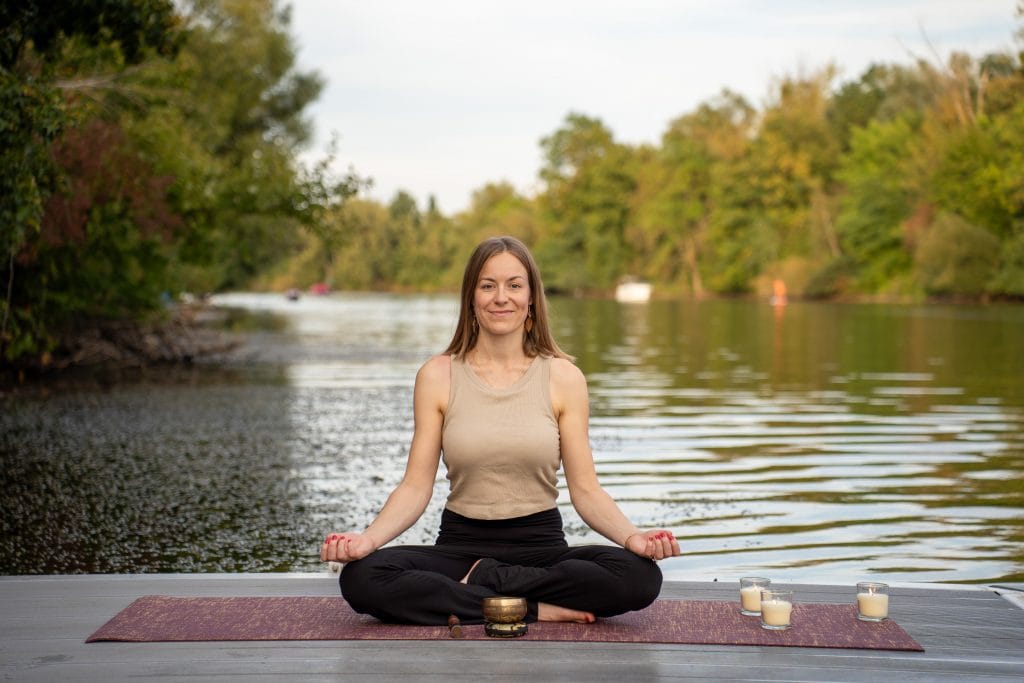 Eine junge Frau meditiert auf einem Steg. Hinter ihr ein See. Auf ihrer Yogamatte sind Kerzen und eine Klangschale.